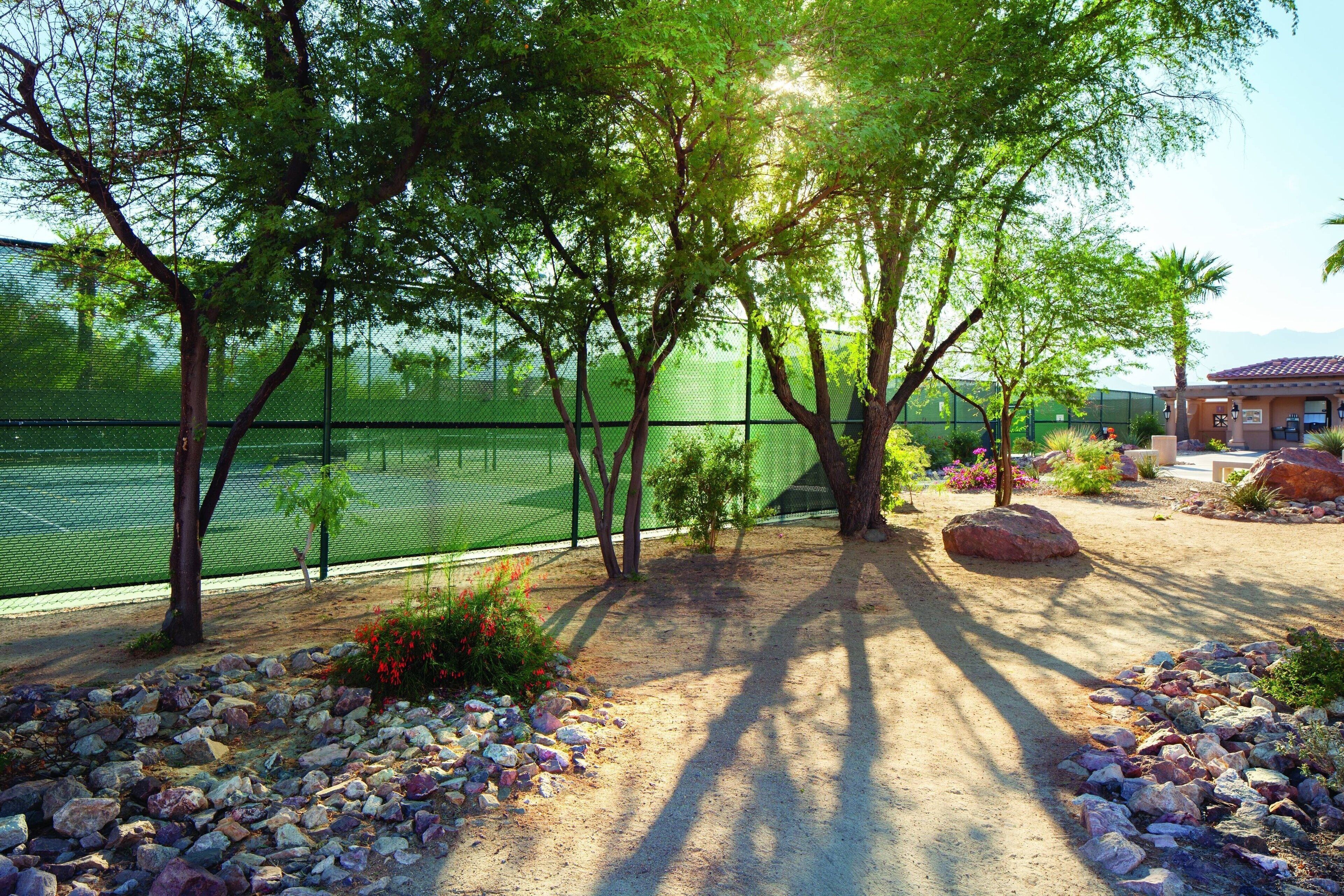 Un court de tennis entouré d’une clôture, avec des arbres et un bâtiment en arrière-plan.