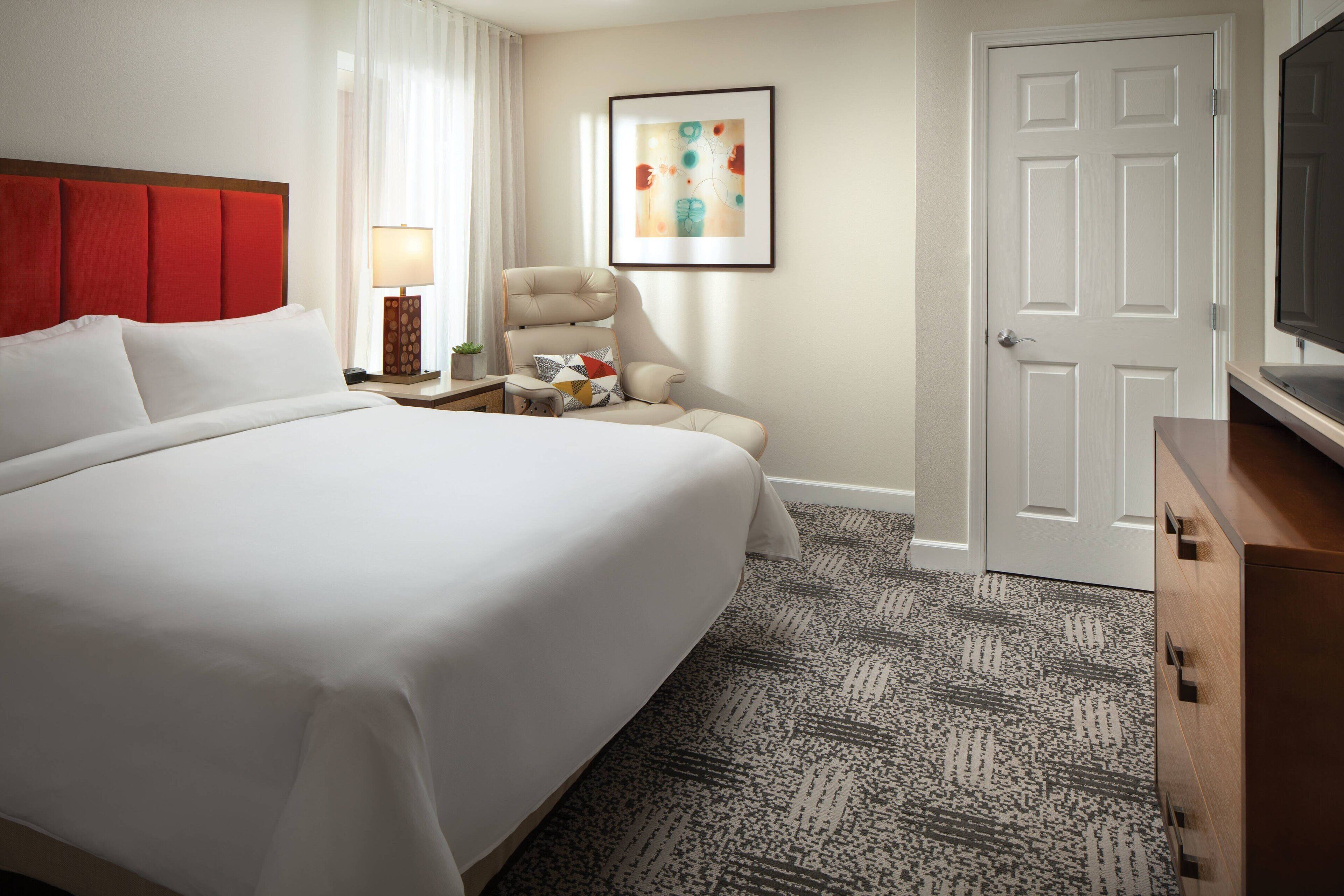 A hotel room with a large bed, a chair, a desk with a lamp, a TV, and a door.