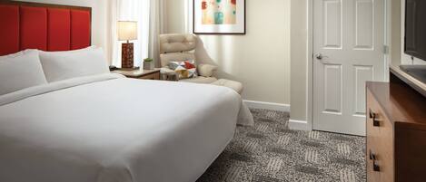 A hotel room with a large bed, a chair, a desk with a lamp, a TV, and a door.