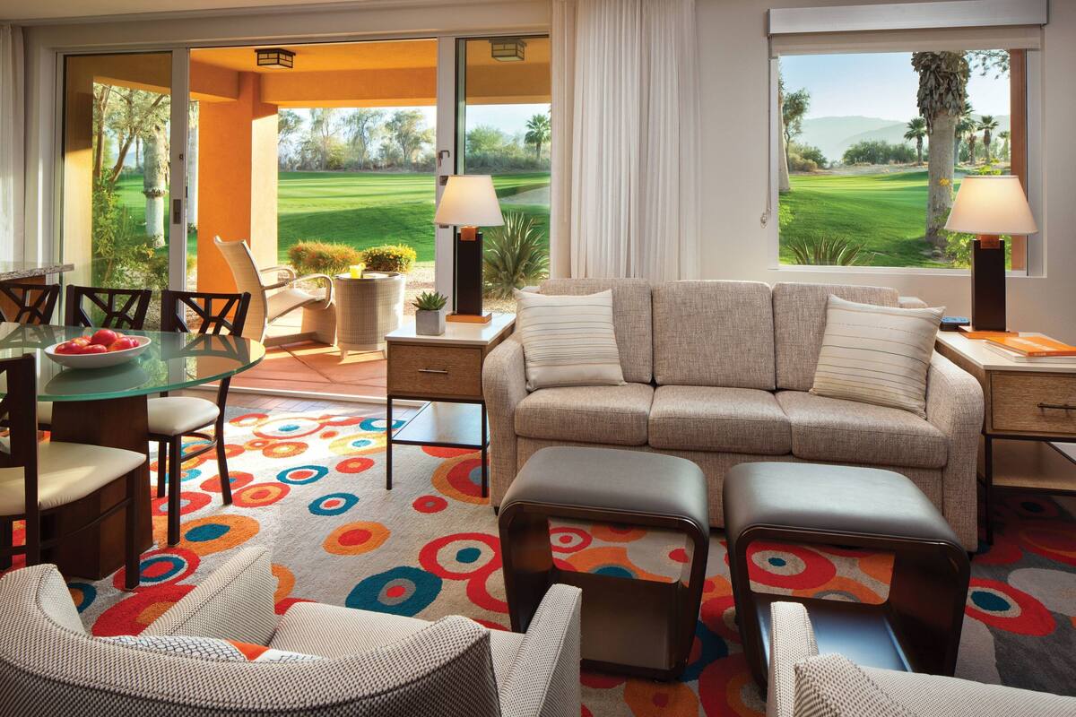 A modern living room with a sofa, ottoman, and chairs, a glass table, and a colorful rug.