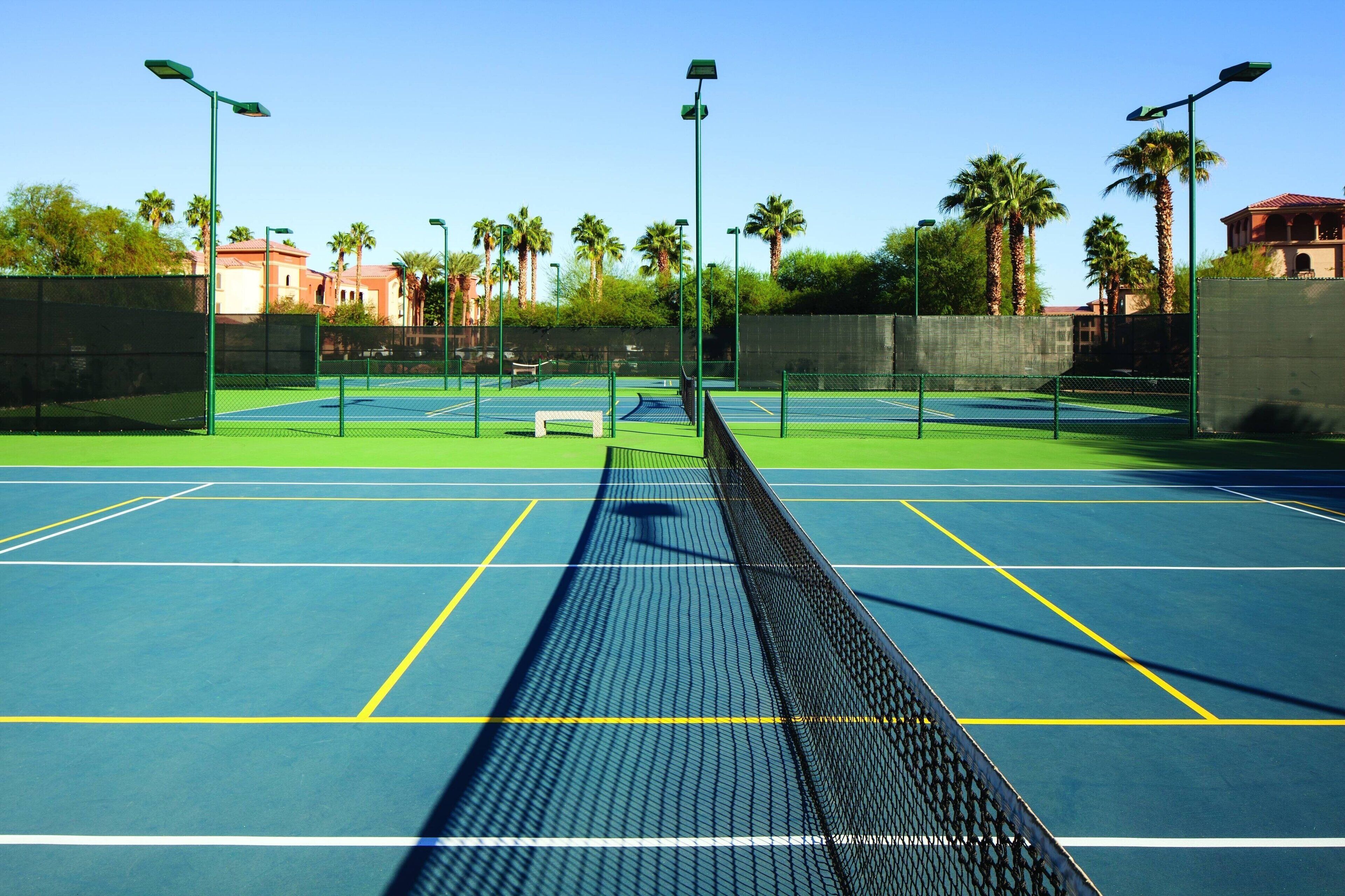 An outdoor tennis court with a net, surrounded by a fence and palm trees.