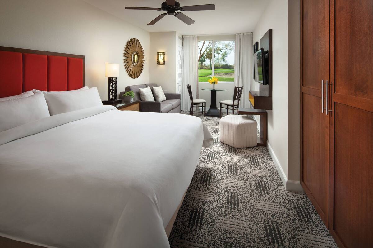 A hotel room with a large bed, a ceiling fan, a seating area with a table and chairs, and a view of a golf course through the window.
