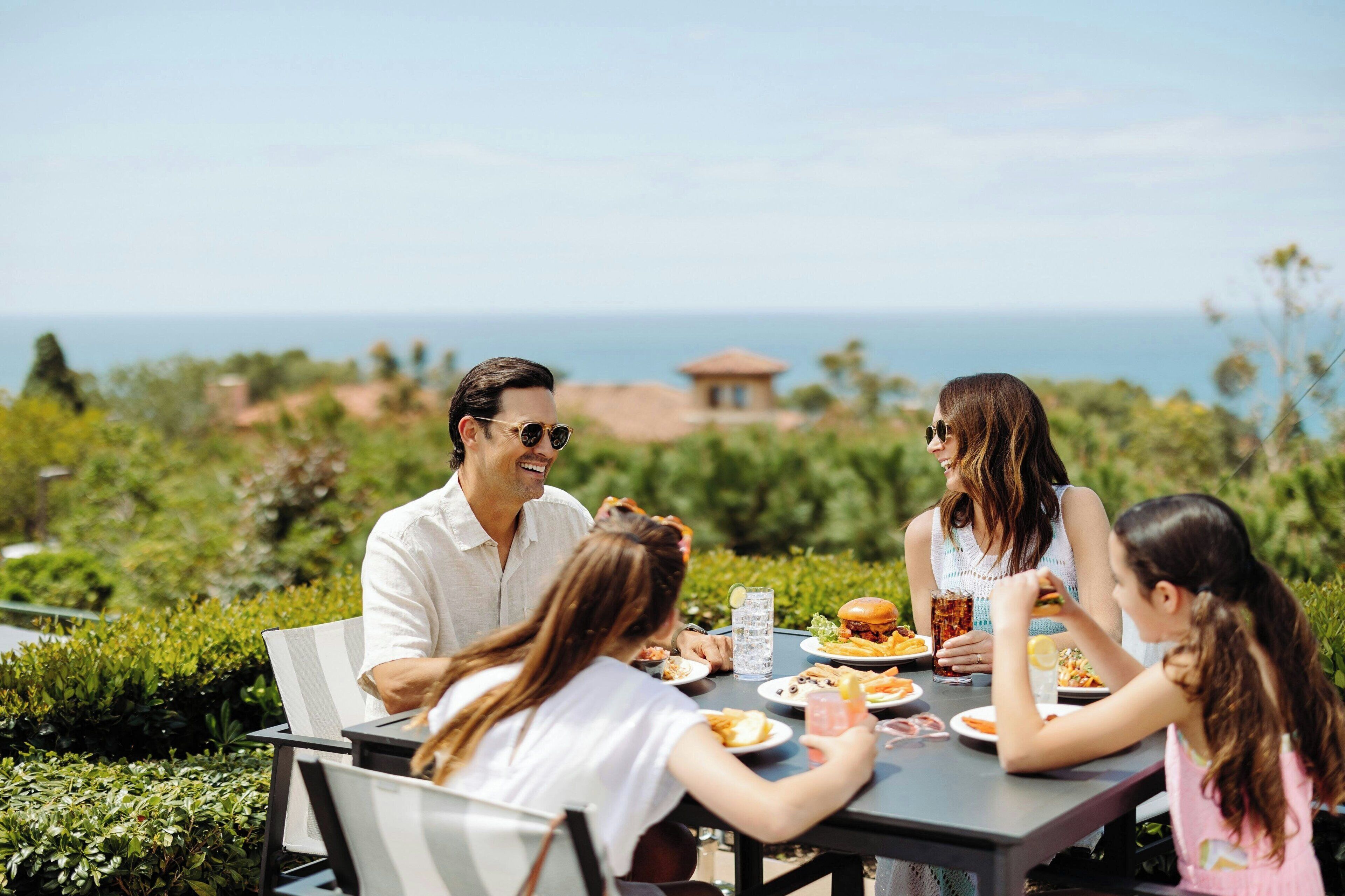 Un grupo de personas cenando en una mesa al aire libre con vistas al océano y vegetación.