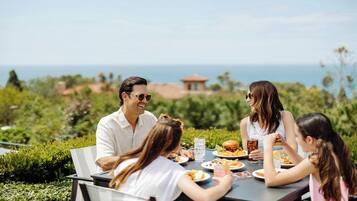 Un grupo de personas cenando en una mesa al aire libre con vistas al océano y vegetación.