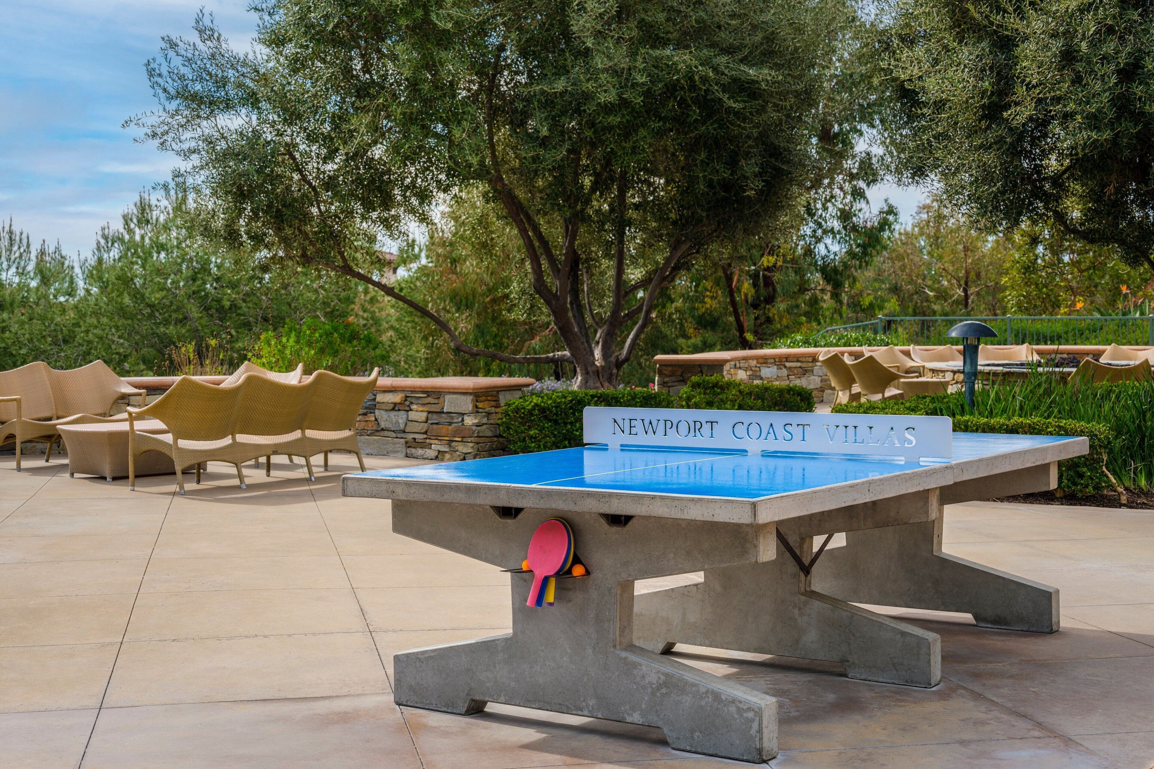 Uma mesa de tênis de mesa com uma placa indicando "NEWPORT COAST VILLAS" em área externa arborizada, com cadeiras e árvores.