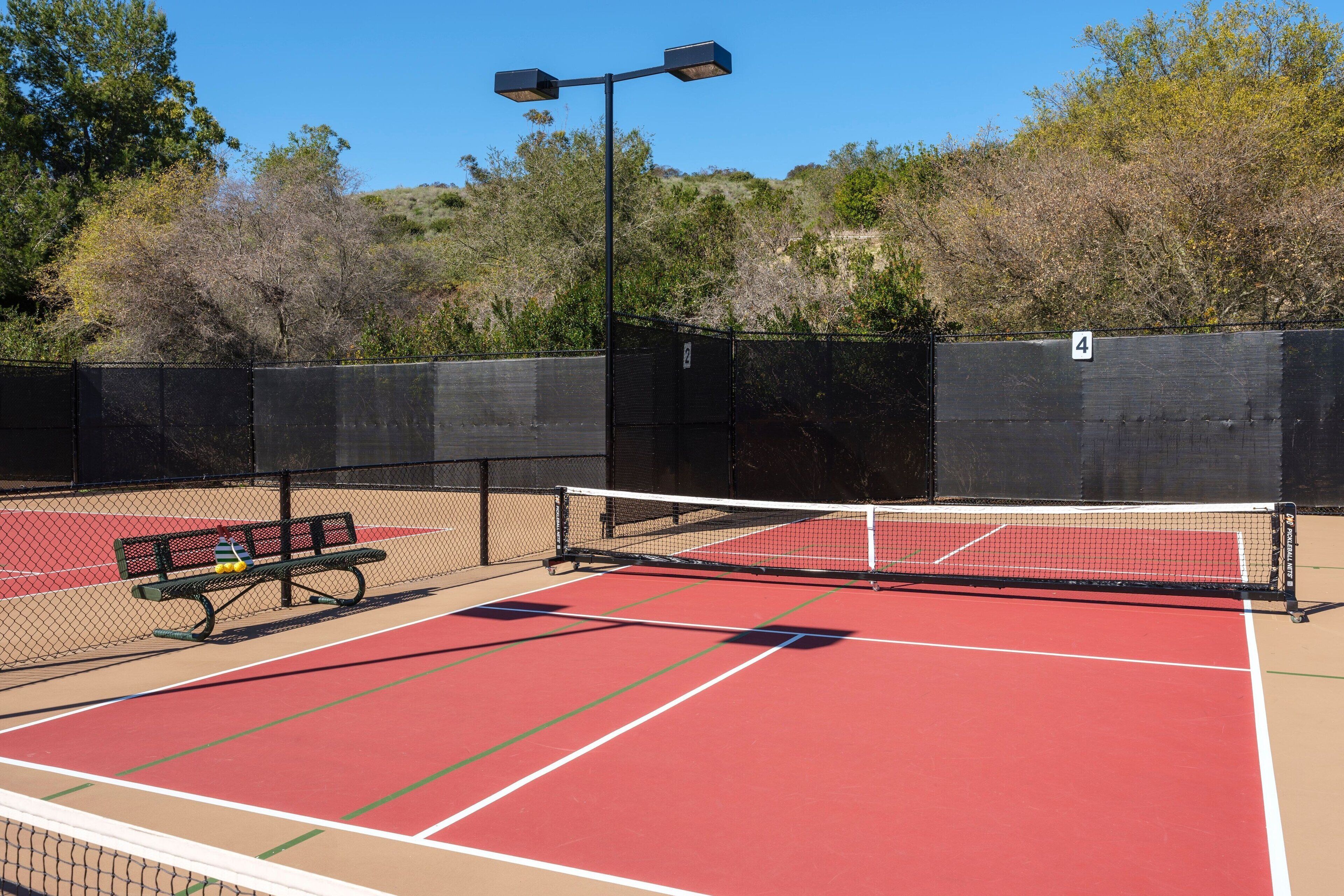 A tennis court with a bench, a fence, and trees in the background.