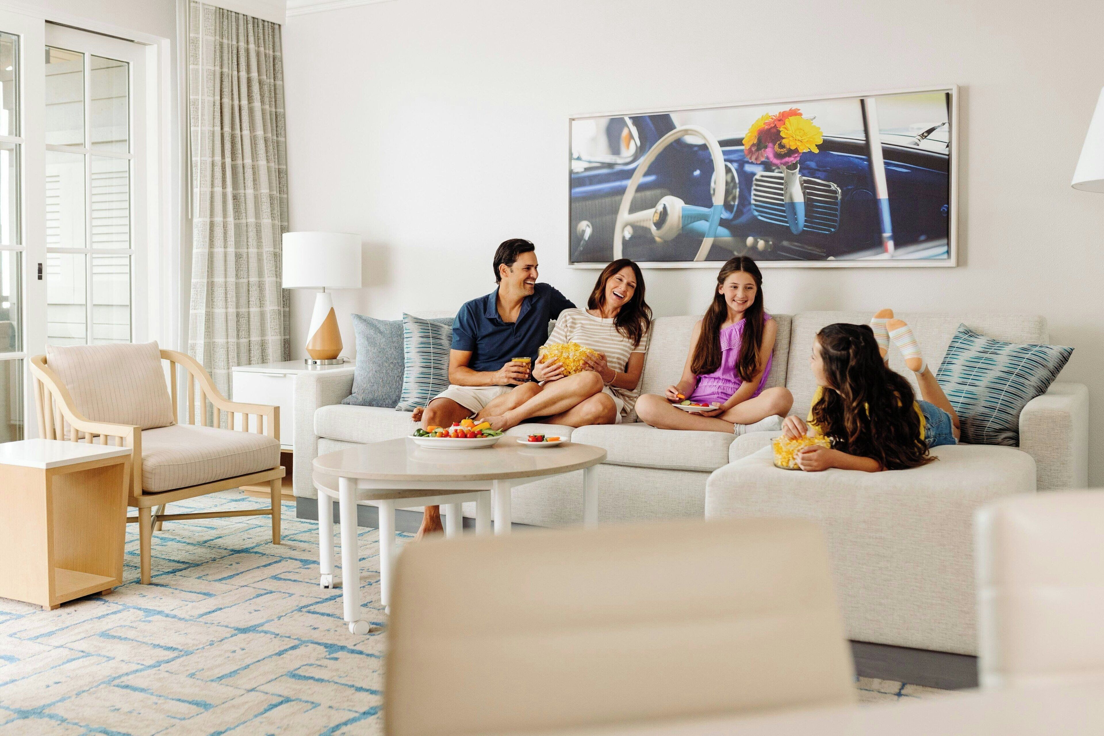 A family sitting on a sofa in a well-lit living room with a modern design, a dining table, and a large window with curtains.