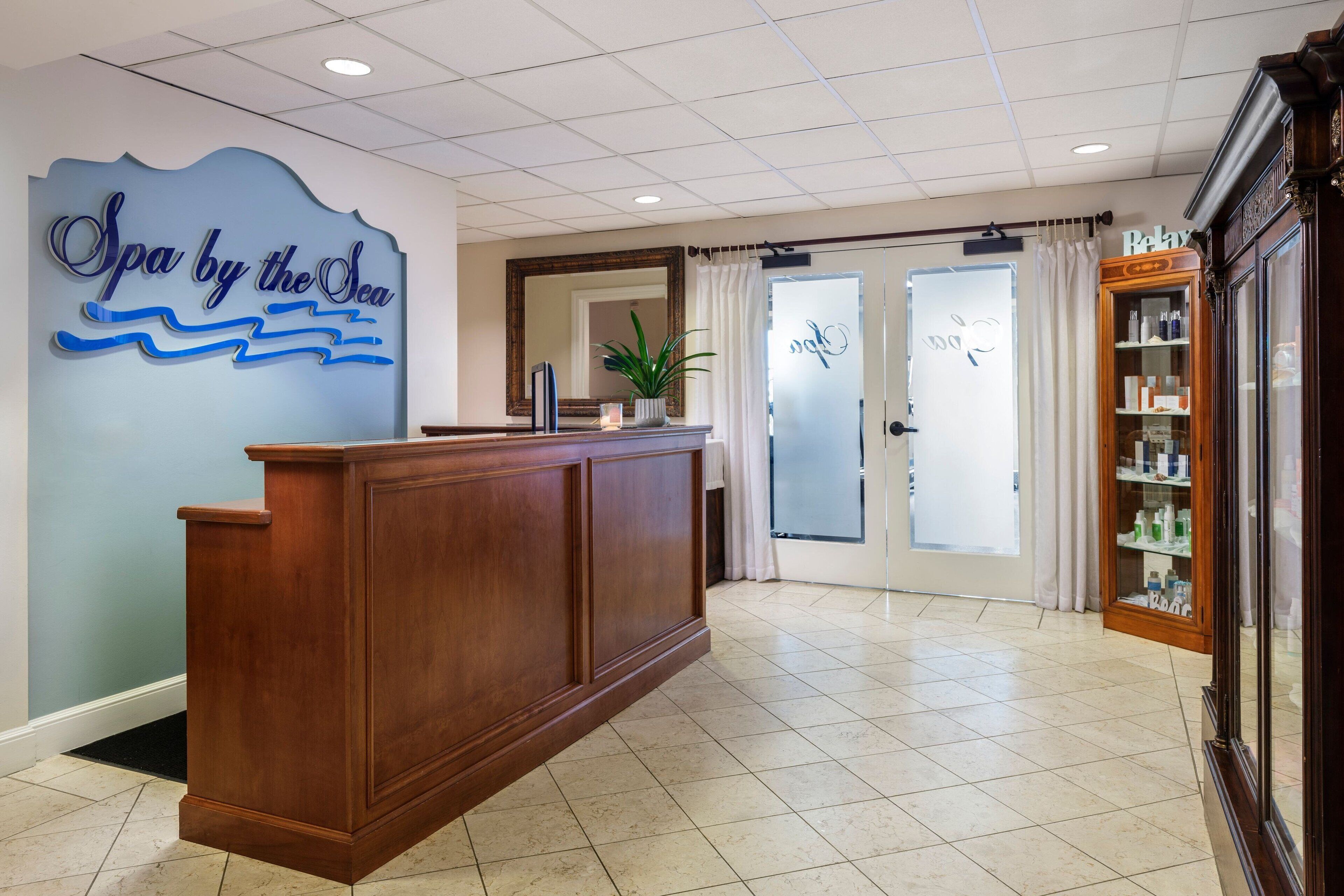 A reception area with a wooden reception desk, a sign reading "Spa by the Sea", and a glass door leading to a room.
