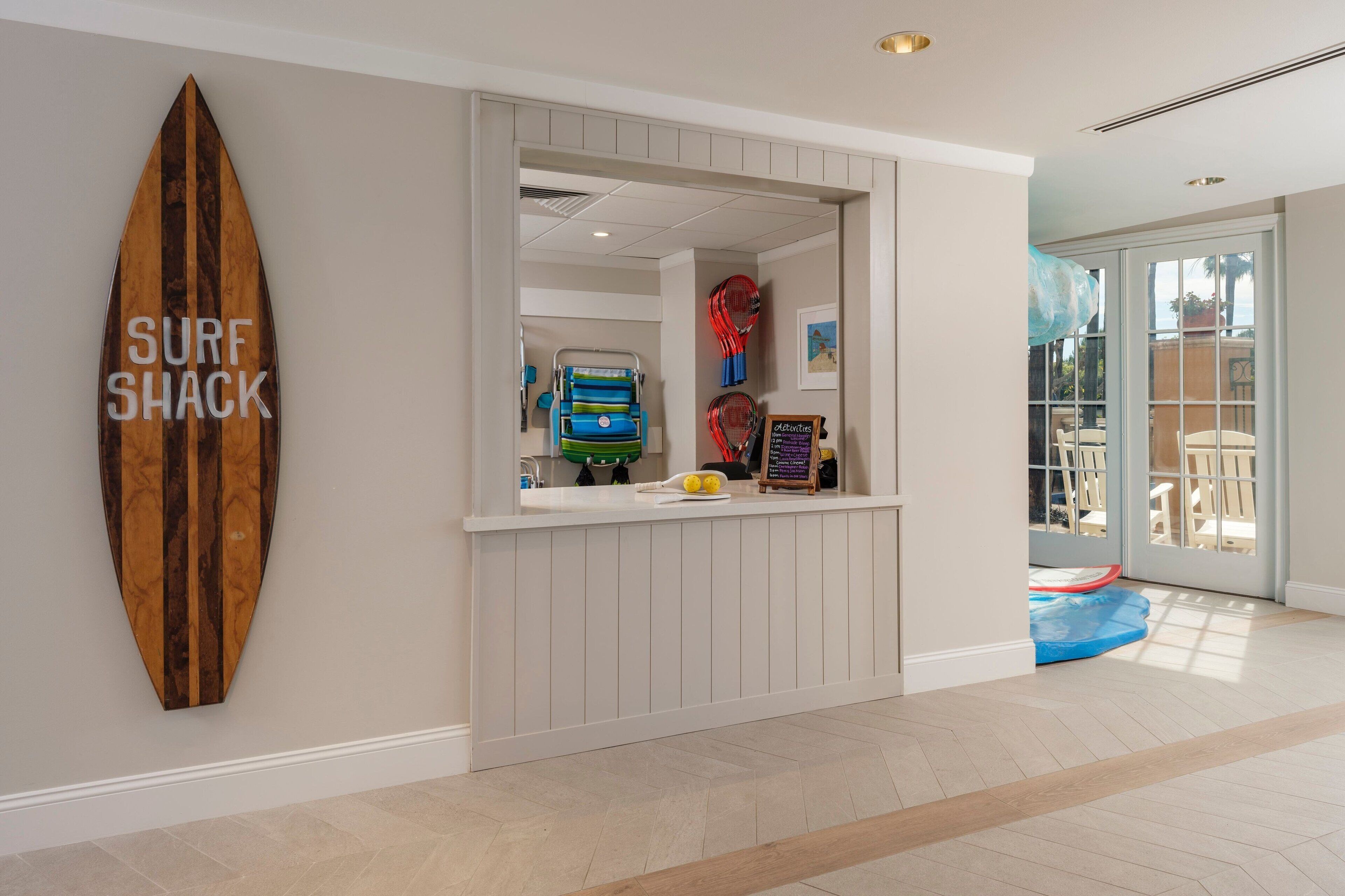 Zona de recepción con un letrero de tabla de surf, mostrador con teléfono y menú, y vista a una piscina exterior.