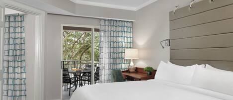 A hotel room with a bed, a ceiling fan, a desk with a lamp, and a view of the outdoors.