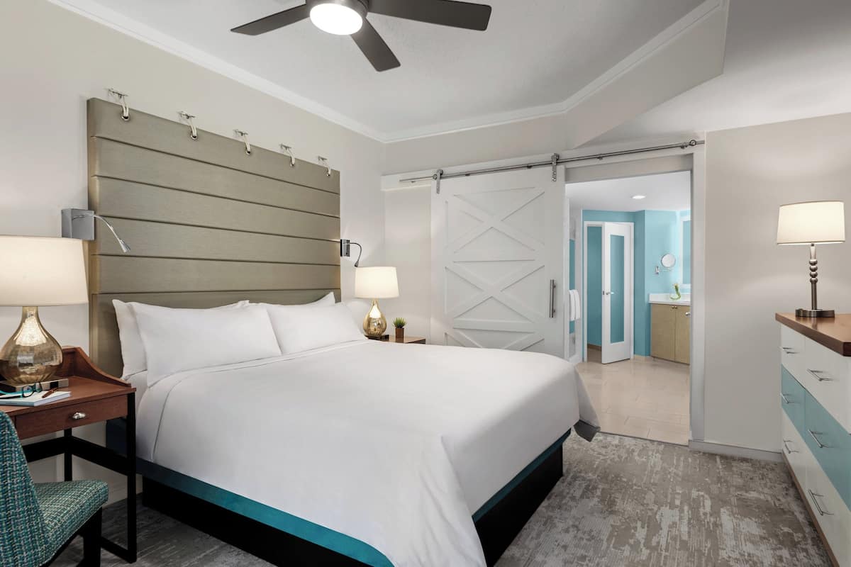 A hotel room with a large bed, a desk with a lamp, a chair, a ceiling fan, and a sliding door leading to a bathroom.