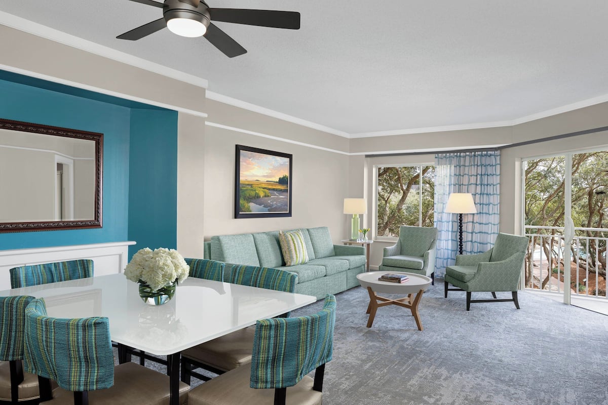A modern living room with a white dining table, teal upholstered chairs, a green sofa, and a ceiling fan.
