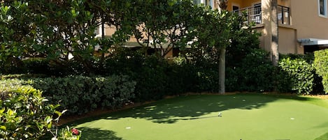 A well-maintained golf course with a putting green, surrounded by lush greenery and a building with balconies.