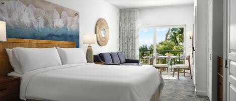 A hotel room with a large bed, a ceiling fan, a painting on the wall, a round mirror, a side table with a lamp, a dark-colored chair, a small table with chairs, and a view of greenery through the window.