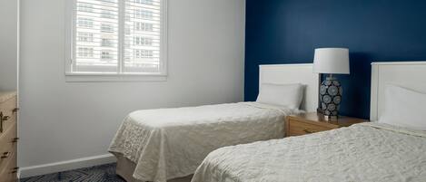 Een slaapkamer met twee bedden, een dressoir, een raam met jaloezieën en een lamp op een nachtkastje.