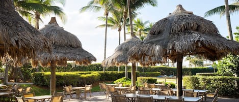 An outdoor dining area with thatched umbrellas, wicker furniture, and palm trees.
