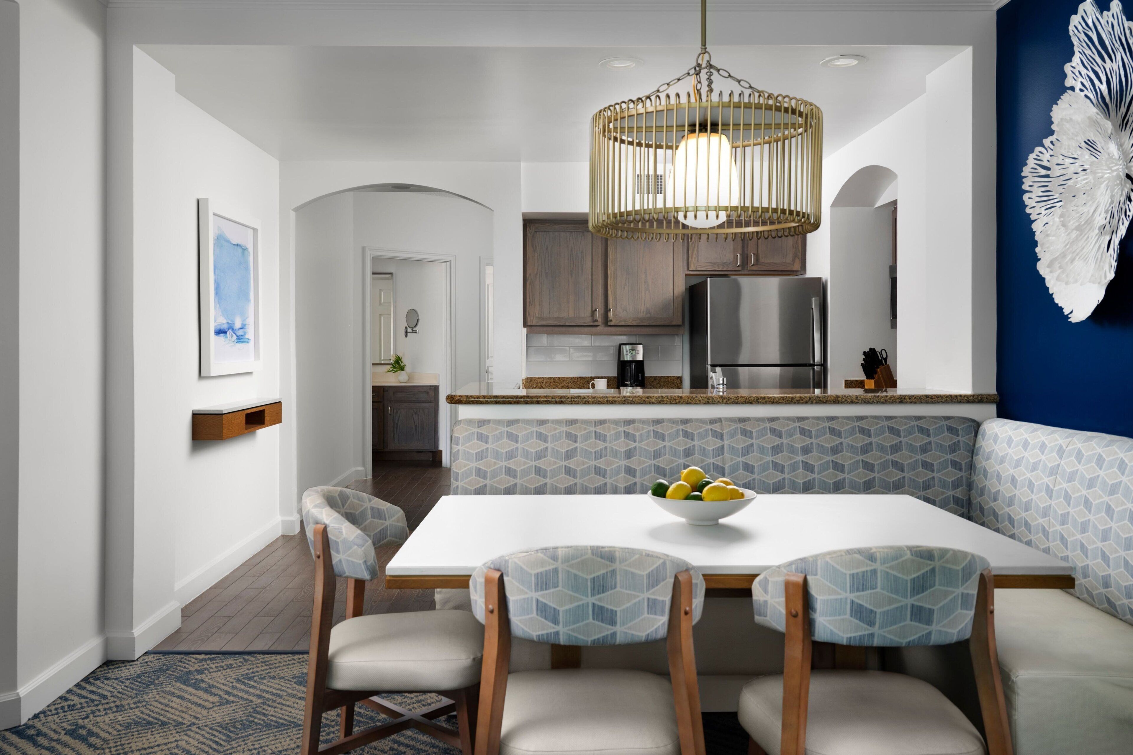 A modern kitchen with a dining area, featuring a white table, blue patterned chairs, and a hanging light fixture.