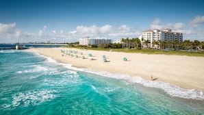 Una playa con aguas turquesas cristalinas, una orilla arenosa y un hotel de varios pisos con balcones con vista al mar.