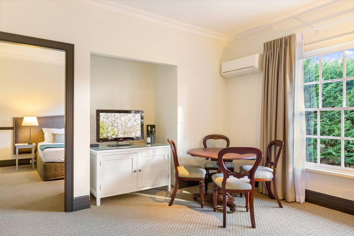 A hotel room with a bed, a desk with a television, a dining table with chairs, and a window with curtains.