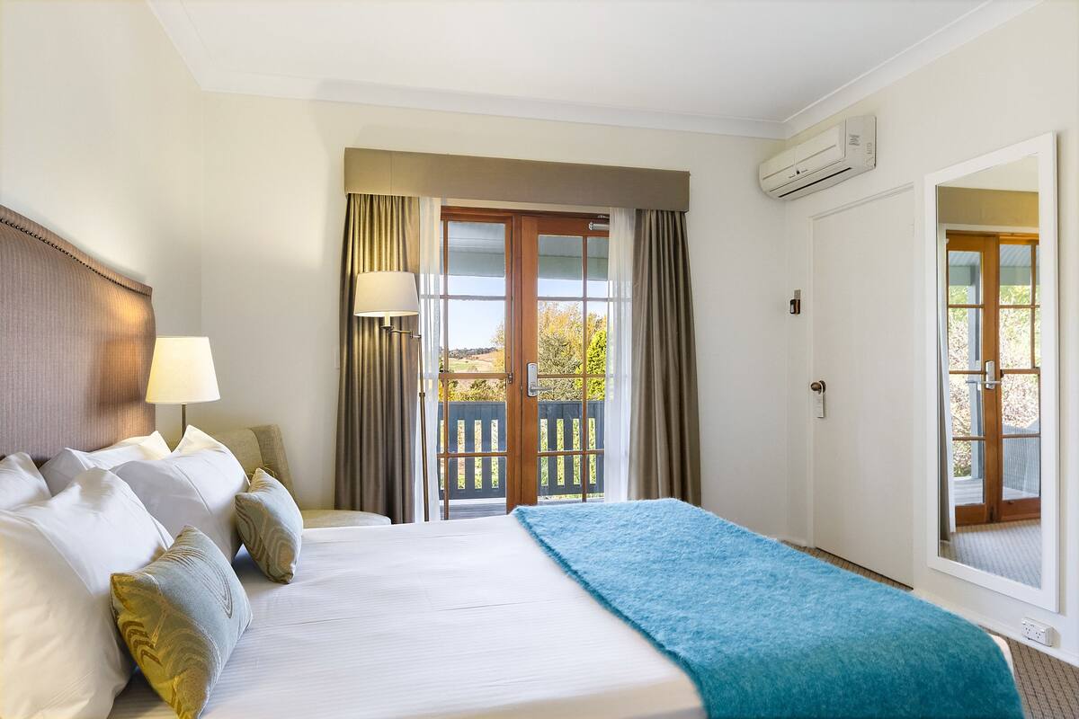 Habitación de hotel con cama, mesita de noche, lámpara, ventana con cortinas y un equipo de aire acondicionado.