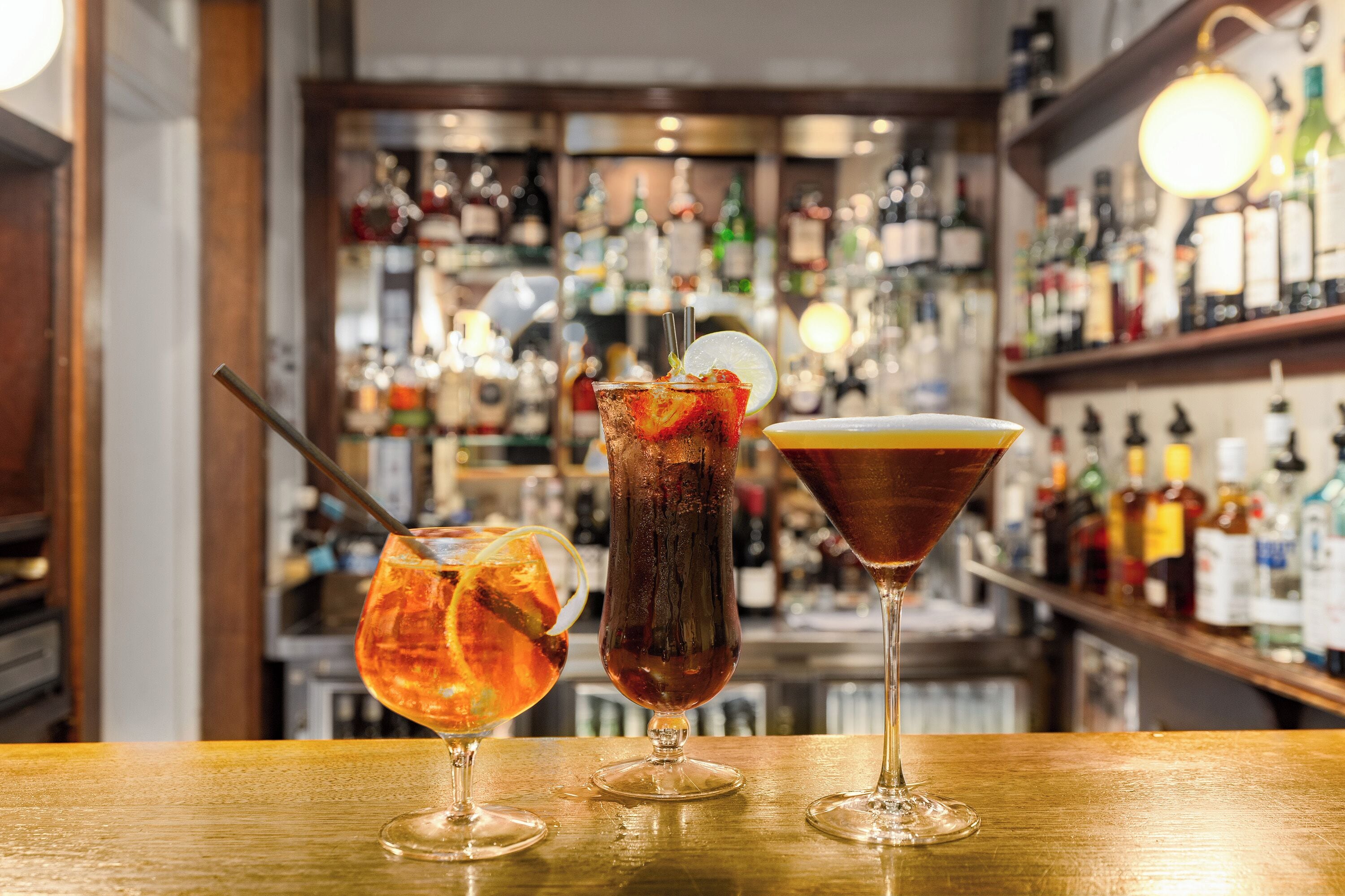 Drei Cocktails auf einer Bar mit unscharfem Hintergrund voller Flaschenregale.