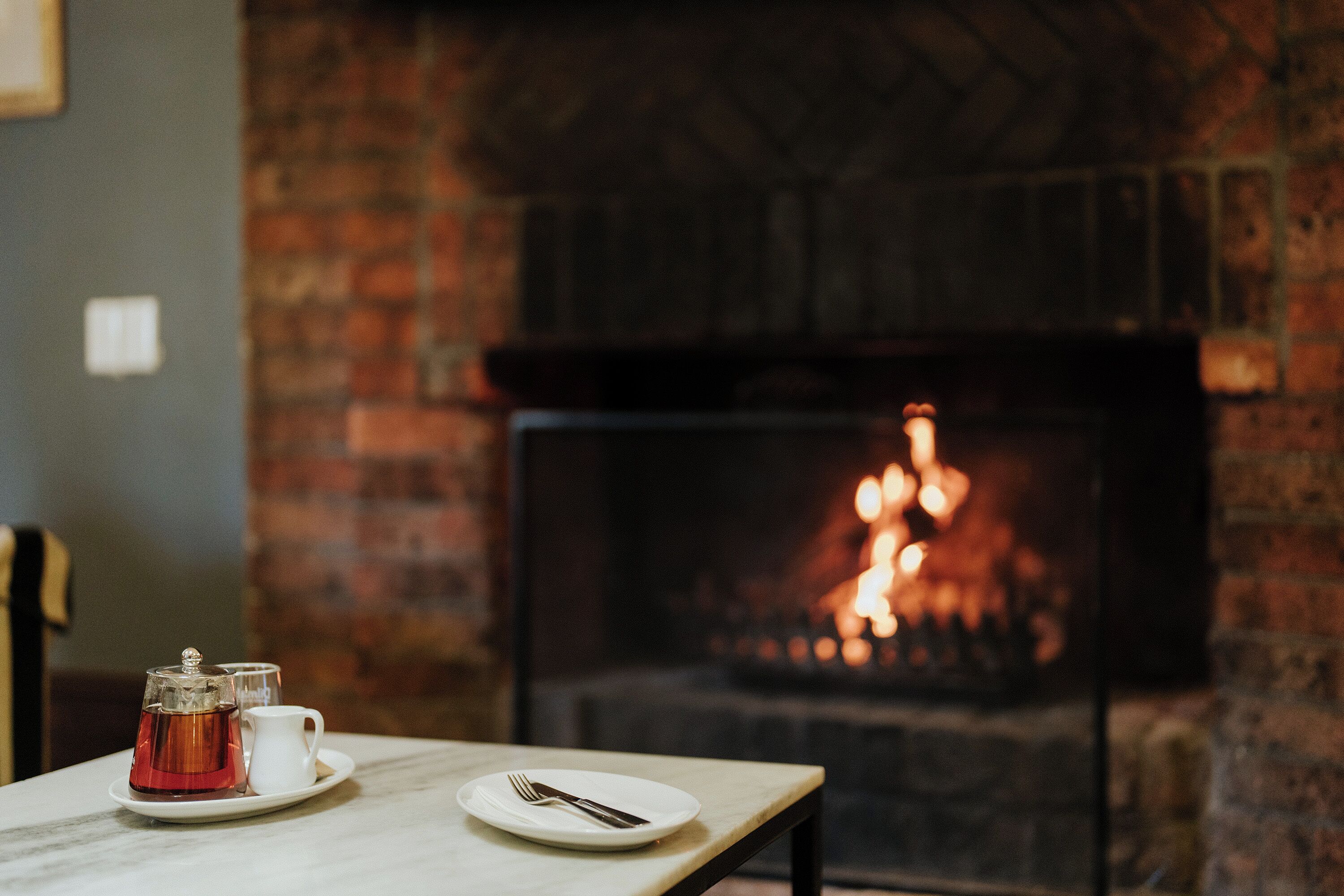 A table with a teapot, cup, and plate, and a fireplace in the background.