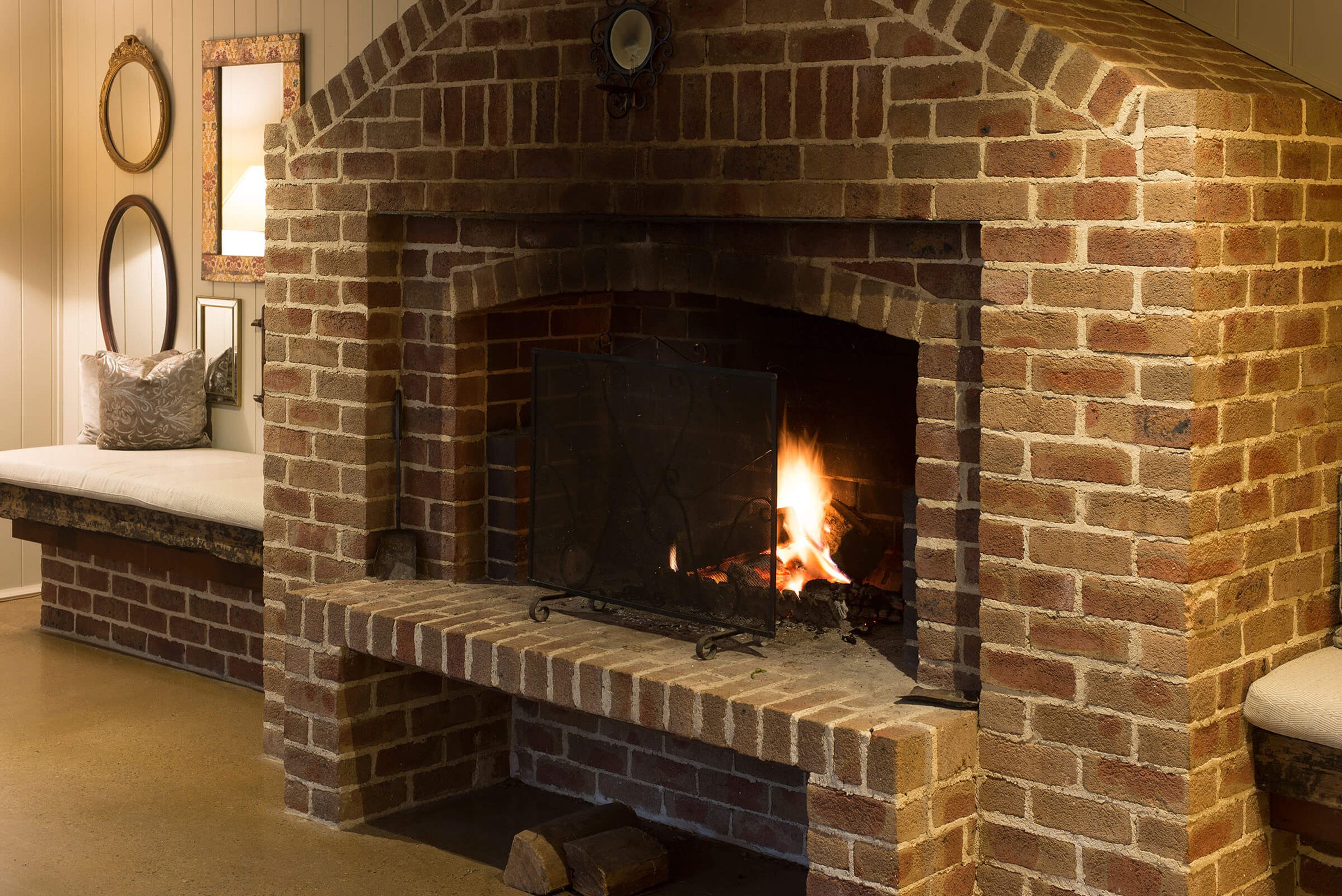 A brick fireplace with a fire burning inside, a clock above it, and a bench with cushions to the left.