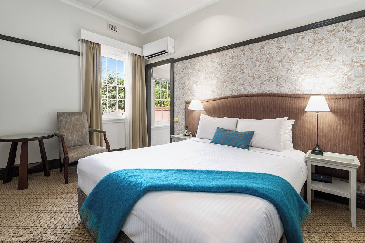 Una habitación de hotel con una cama grande, dos mesitas de noche con lámparas, una silla y un ventanal con cortinas.