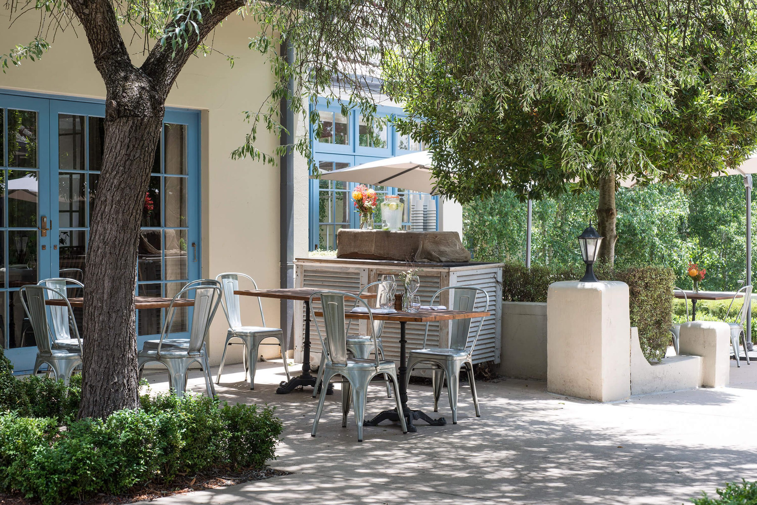 Un patio al aire libre con sillas y una mesa de metal, rodeado de vegetación y un edificio con puertas azules.