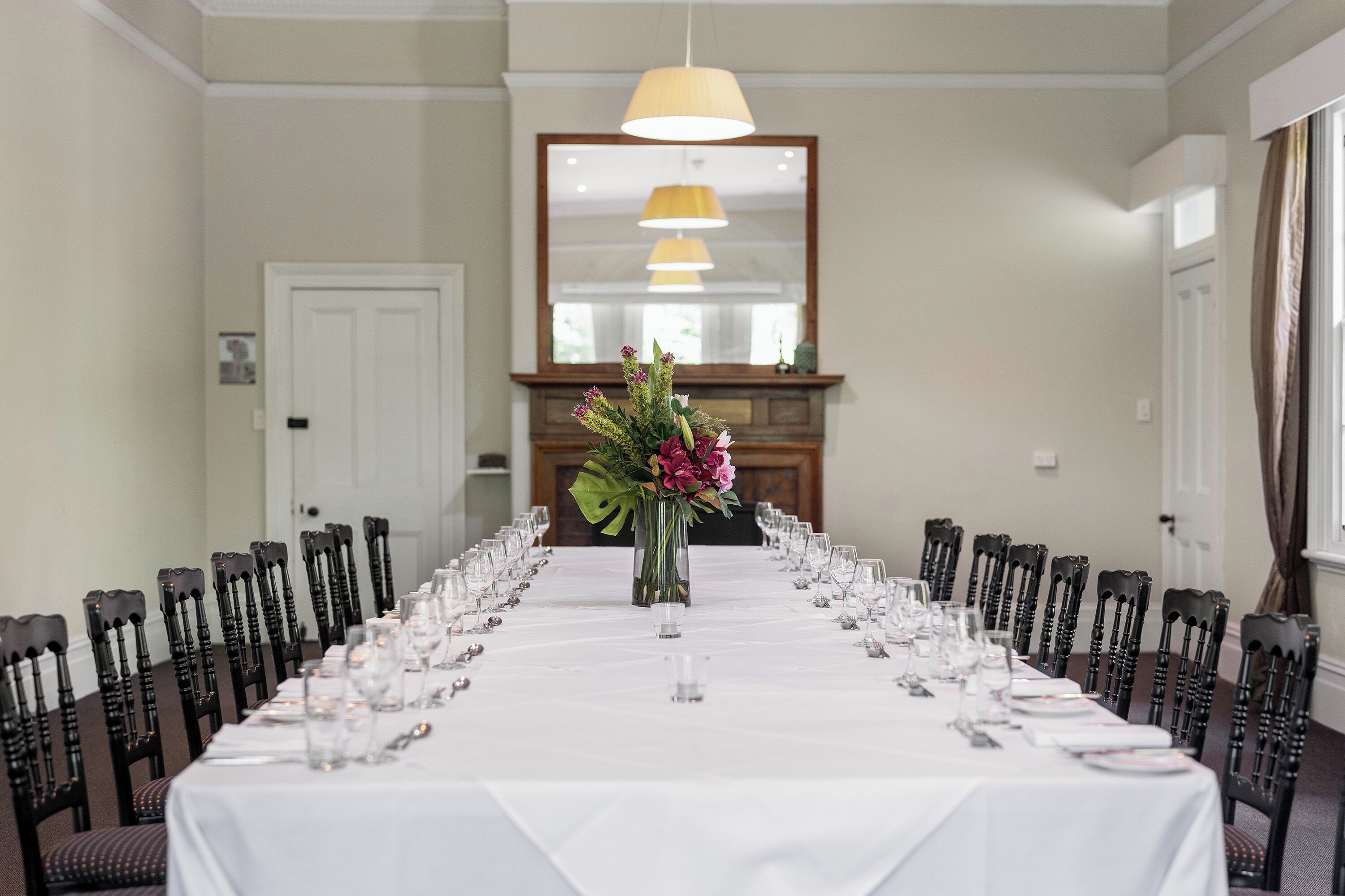 Una larga mesa de comedor dispuesta para una comida formal, con sillas a ambos lados, un jarrón de flores en el centro y un espejo sobre la mesa.