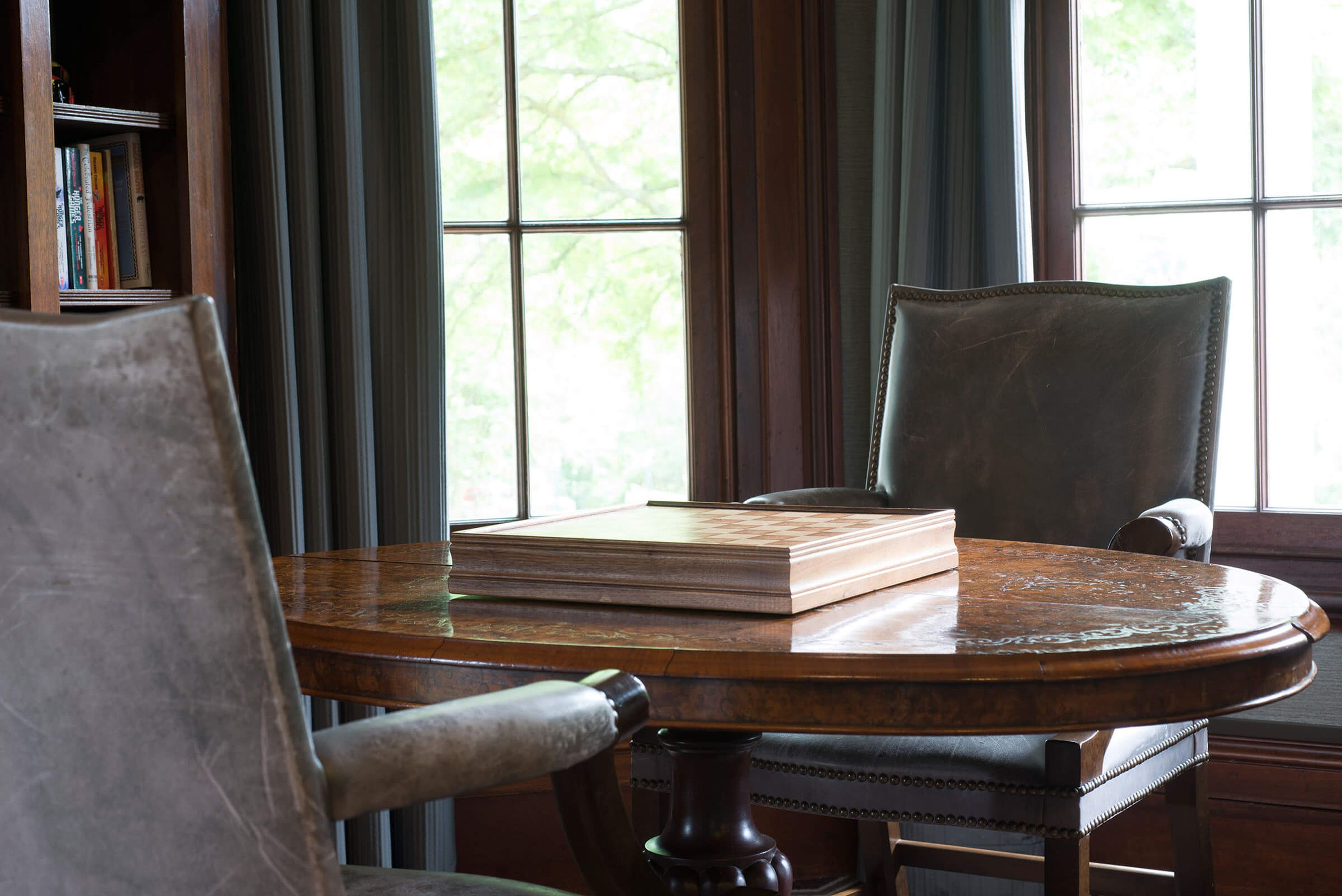 Une table ronde en bois, sur laquelle repose un livre, deux chaises, une bibliothèque et une fenêtre avec des rideaux.