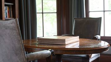 Une table ronde en bois, sur laquelle repose un livre, deux chaises, une bibliothèque et une fenêtre avec des rideaux.