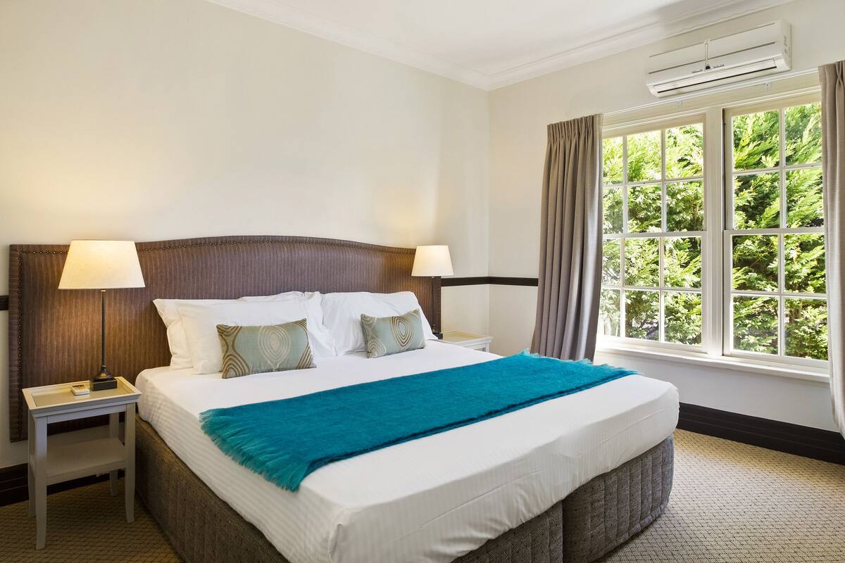 Una cama bien hecha con sábanas blancas y una manta verde azulado, una mesita de noche con una lámpara, y una ventana con cortinas.