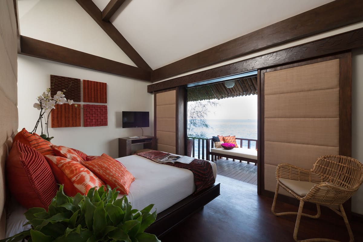 A bedroom with a bed, a TV, a balcony with a wicker chair, and a view of the sea.