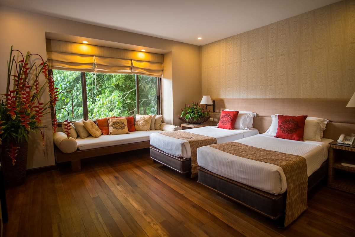 A hotel room with two beds, a sofa, a nightstand, and a large window with a view of greenery.