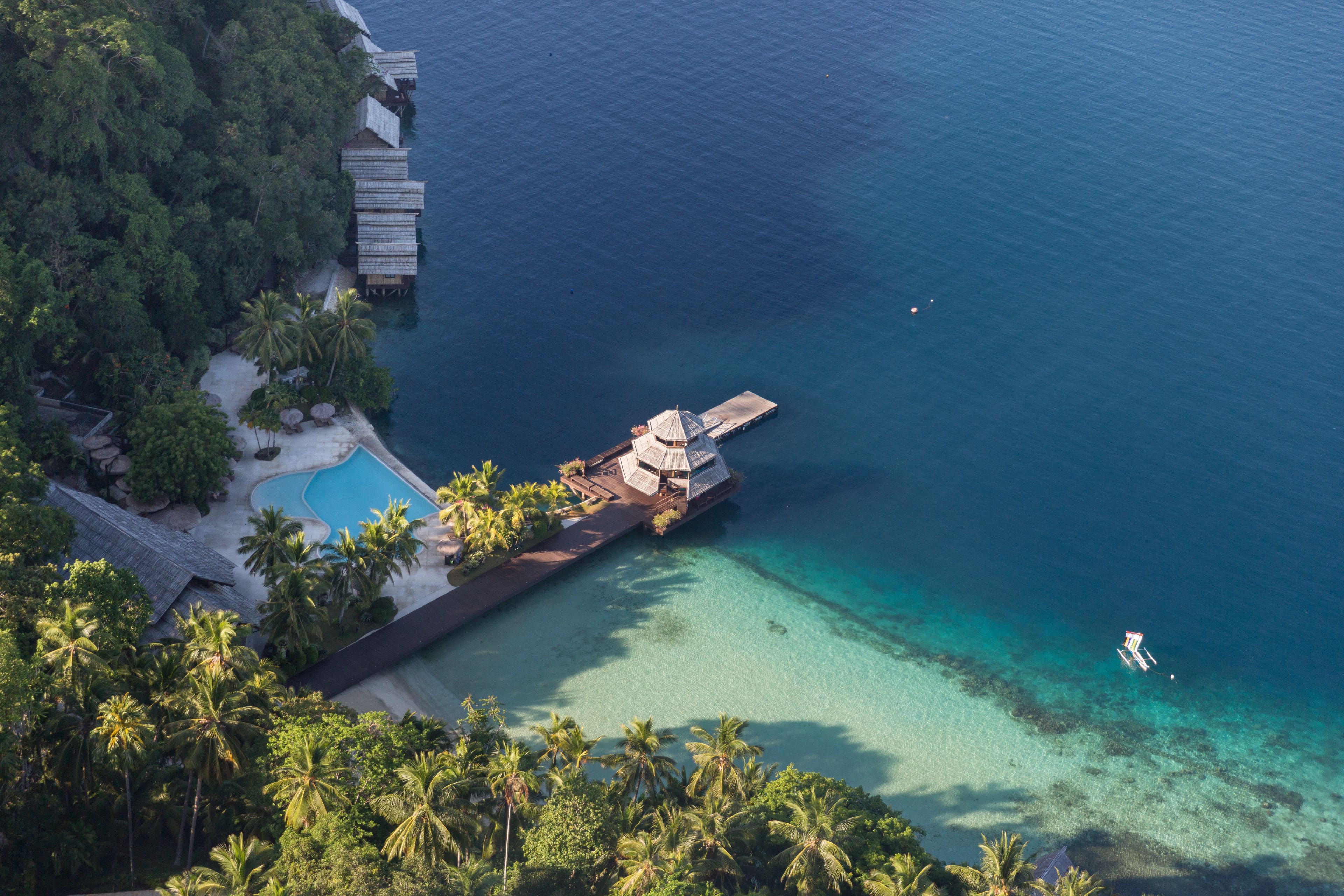 An aerial view of a resort with a swimming pool, palm trees, and a pier extending into the ocean.