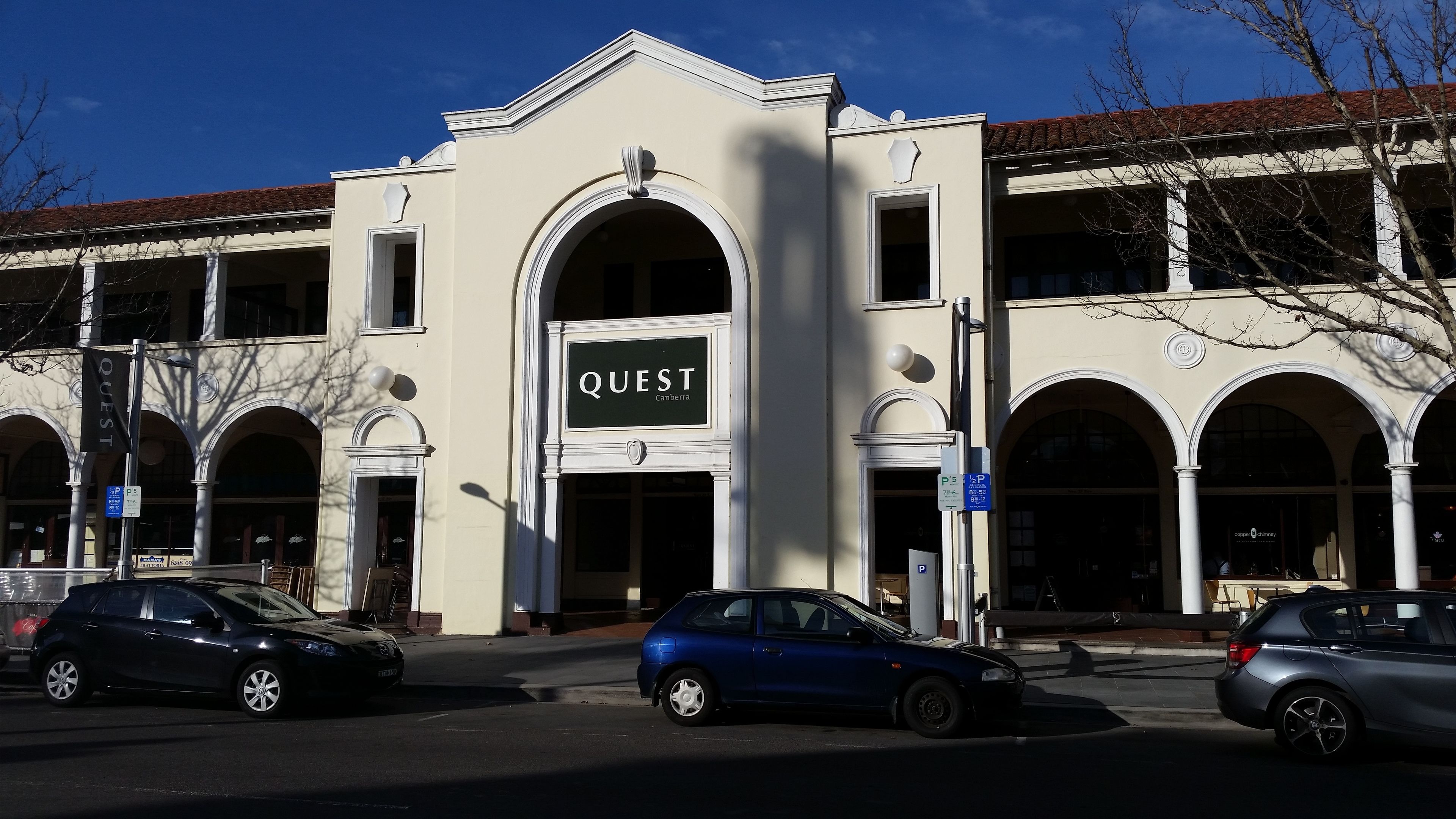 A building with the sign "QUEST Canberra" above the entrance. There are cars parked in front.