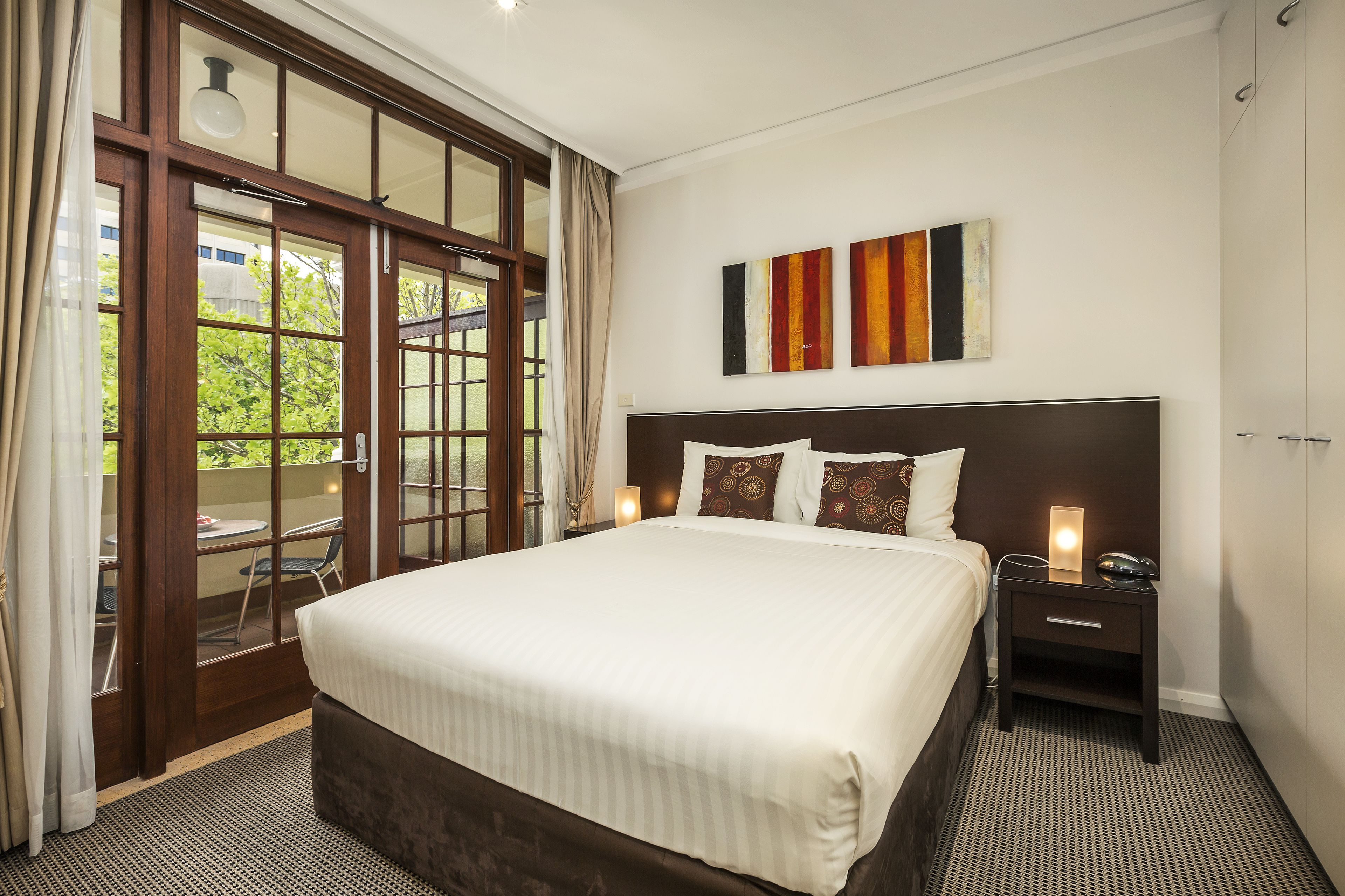 A hotel room with a large bed, a nightstand, a wall-mounted artwork, and a sliding glass door leading to an outdoor area.