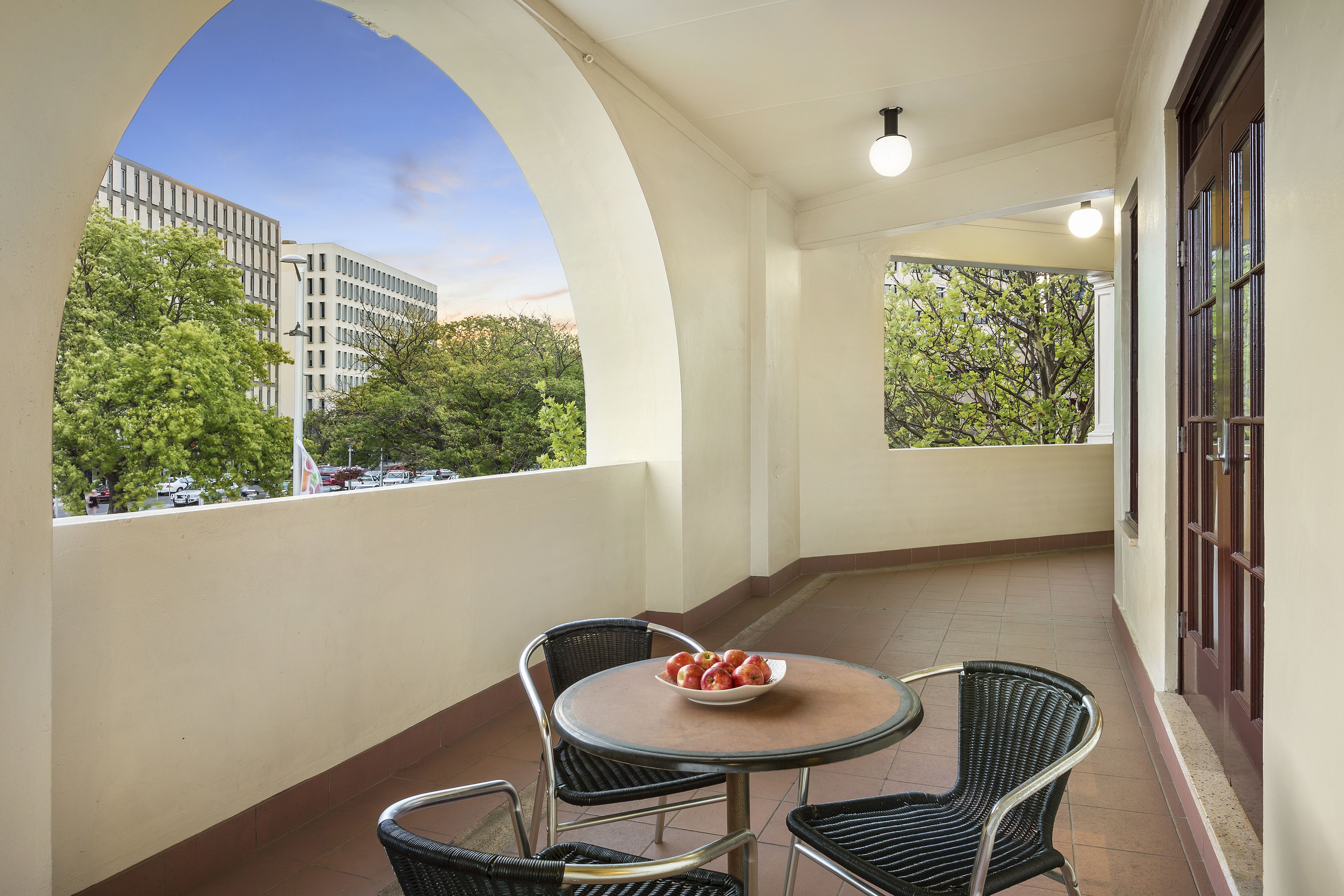 A balcony with a round table and chairs, a bowl of fruit, and a view of buildings and trees.