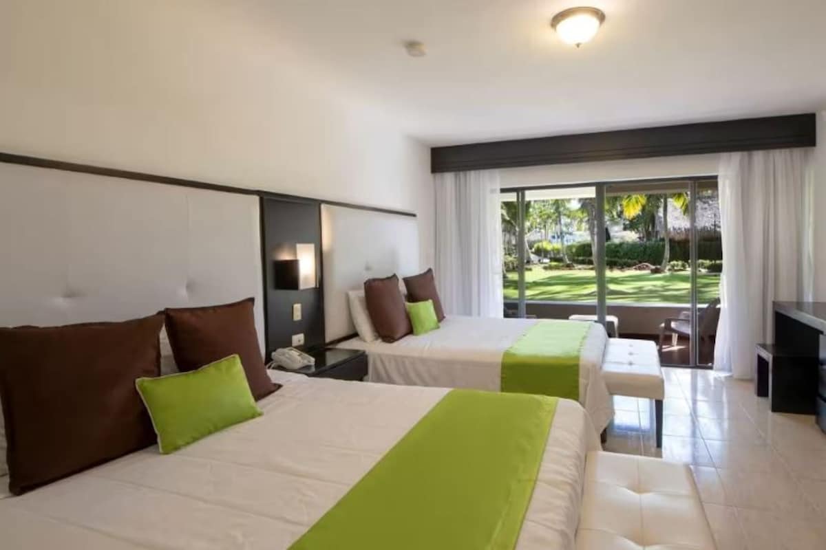 A hotel room with two beds, a desk, and a view of a garden.