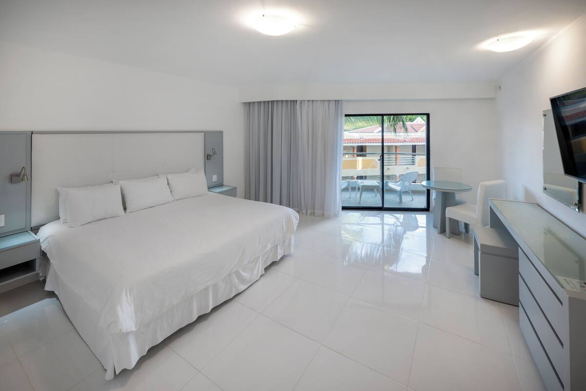 A modern hotel room with a large bed, a small desk, a TV, and a balcony with a view.