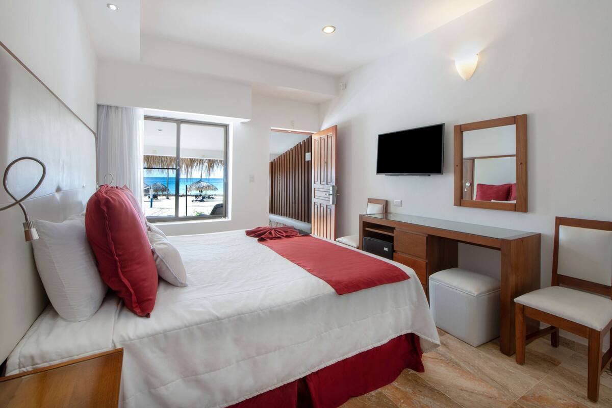 A modern hotel room with a large bed, a desk, a chair, a TV, and a view of the beach.