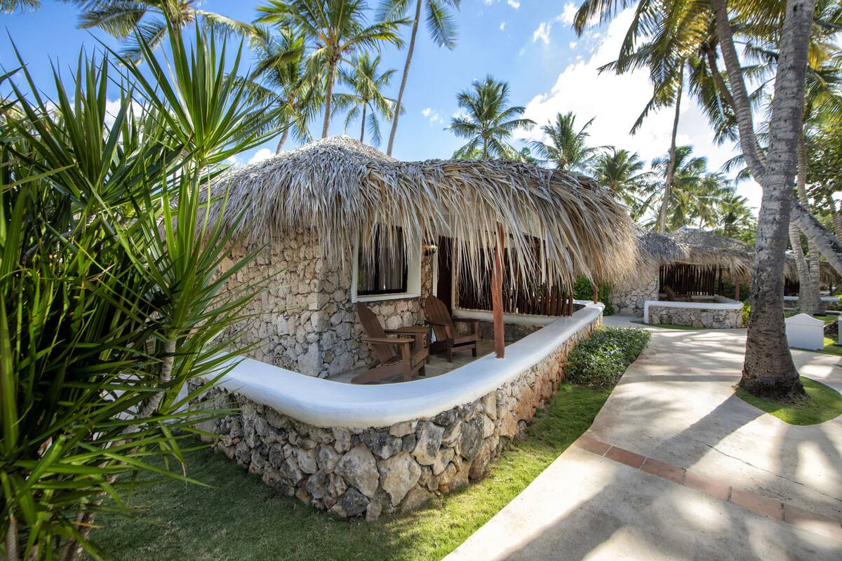 A thatched-roof hut with a stone wall, a seating area with chairs, and a pathway leading to another hut.