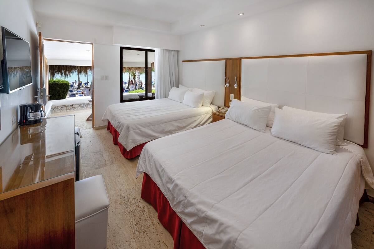 A hotel room with two beds, a TV, a desk, and a view of the beach.