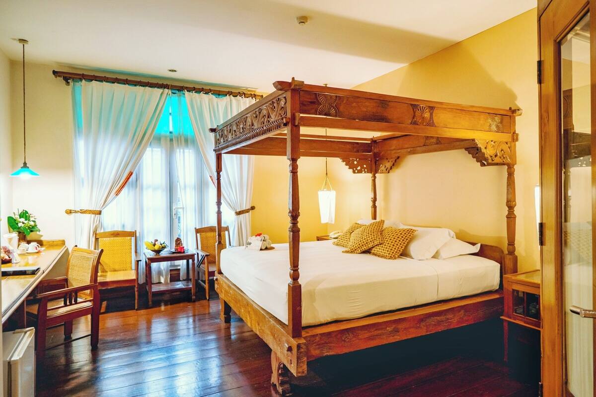 Ein Bett mit Baldachin mit Holzrahmen, ein Essbereich mit Tisch und Stühlen sowie ein Blick ins Freie durch ein Fenster mit durchsichtigen Vorhängen.