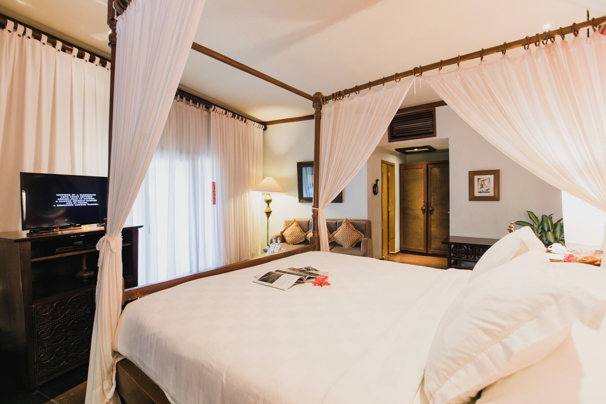 A four-poster bed with white linens, a television on a wooden stand, a bedside table with a lamp, and a mirror on the wall.