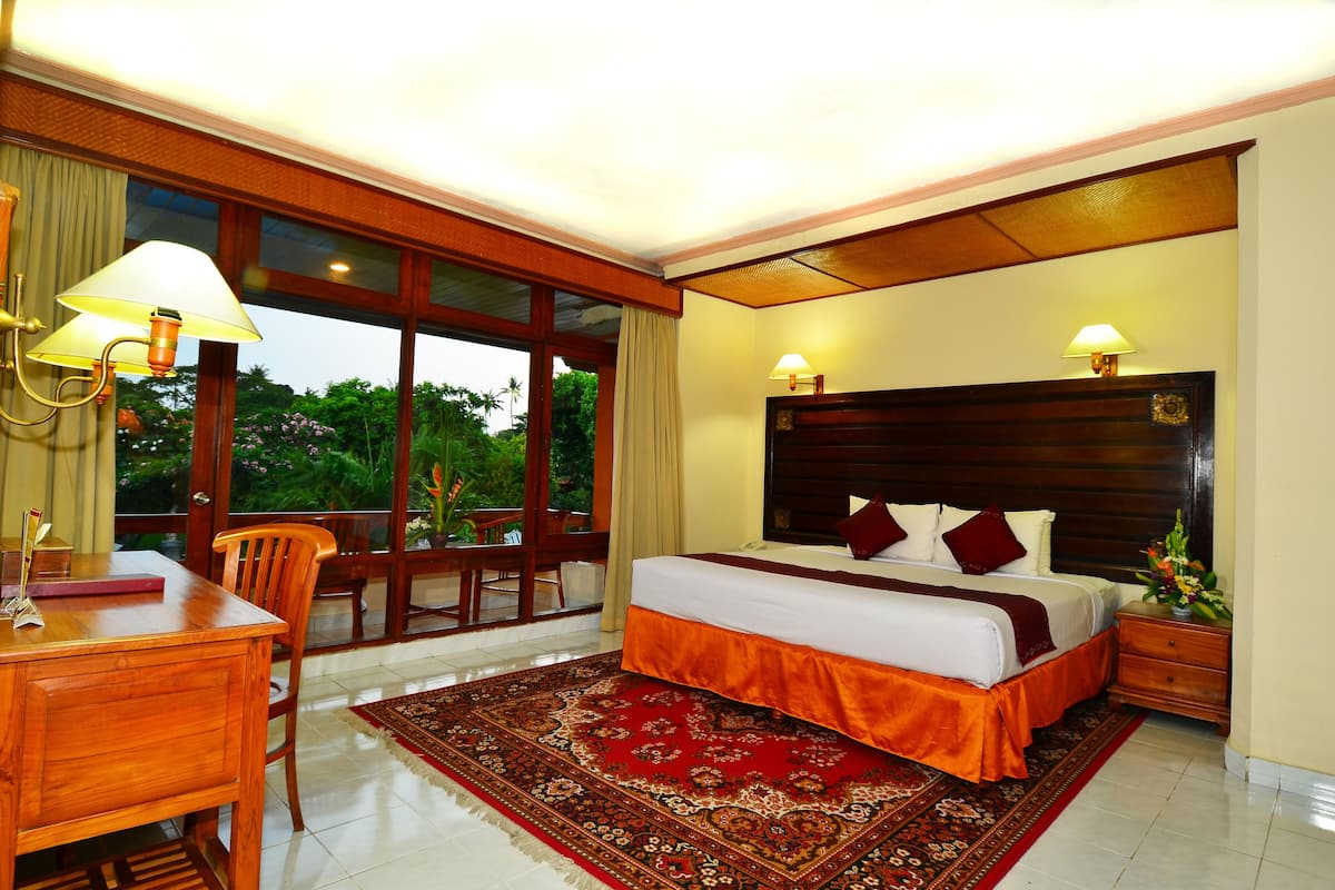 A hotel room with a large bed, a desk, and a chair. There is a window with a view of greenery outside.
