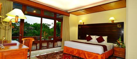 Habitación de hotel con una cama grande, dos lámparas de noche, un escritorio de madera, una silla, un tapete con estampado rojo y vistas a zonas verdes a través de las ventanas.