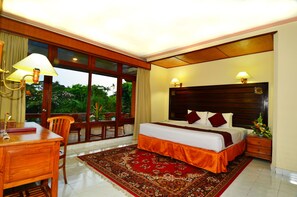 A hotel room with a large bed, two bedside lamps, a wooden desk, a chair, a red patterned rug, and a view of greenery through the windows.