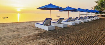 Una playa con filas de sillas de playa blancas y sombrillas azules, un barco en el agua y un atardecer de fondo.